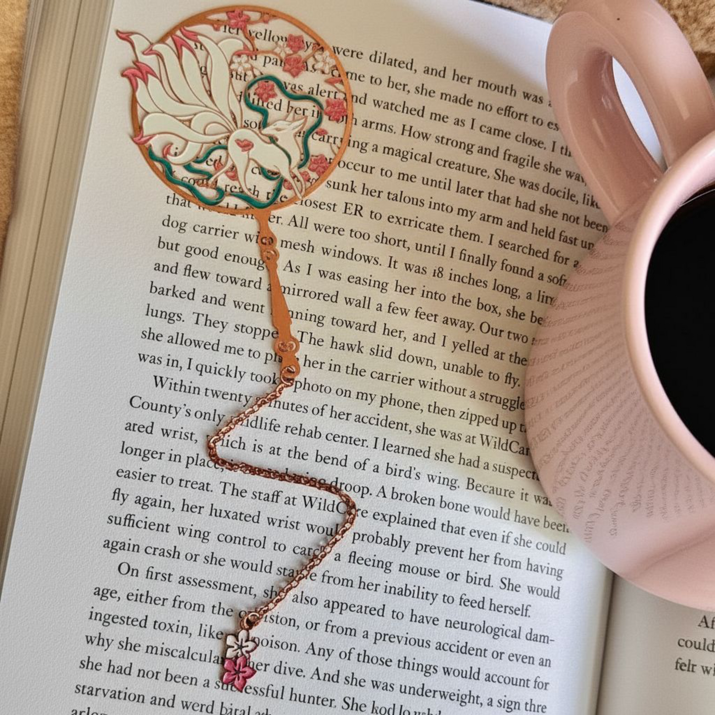 open book with nine tailed fox brass metal bookmark that has a chain and a sakura charm on the end lying next to a pink coffee mug
