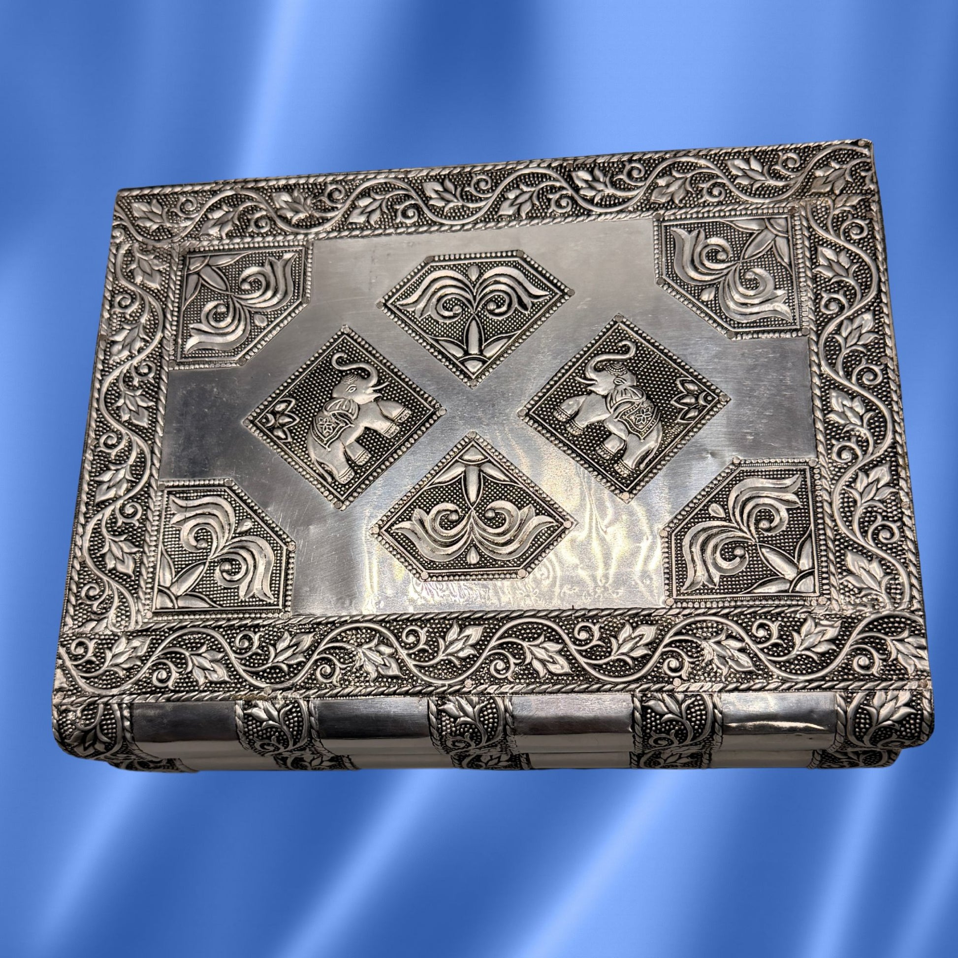 An antique rectangular silver jewelry metal box with intricate embossed floral borders and four central diamond motifs featuring elephant carvings, shown against a blue gradient background.