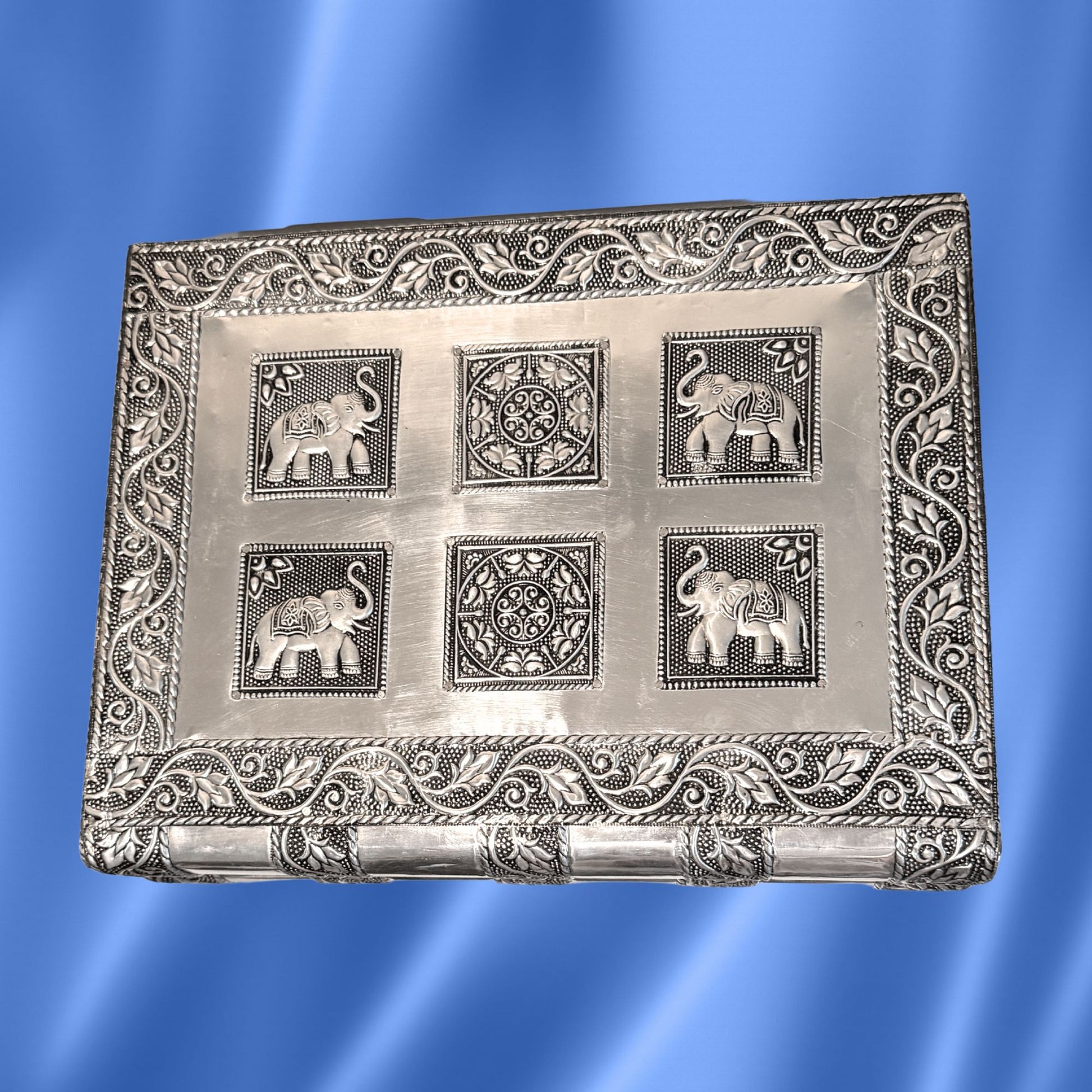An Indian rectangular silver-toned decorative jewelry box with embossed floral borders and four raised elephant and geometric medallion motifs on the front, shown against a blue satin-like background.
