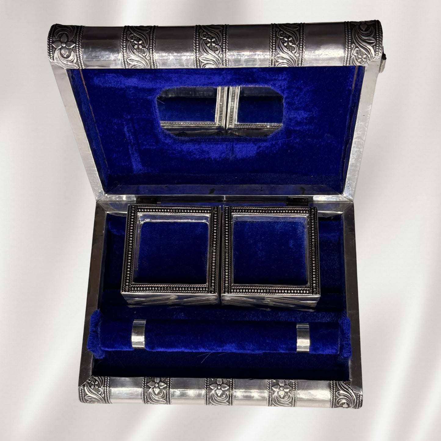 A vintage silver-toned jewelry box with a deep blue velvet interior, shown open to display a matching pair of rectangular trays and a cylindrical dowel to hold bracelets inside.