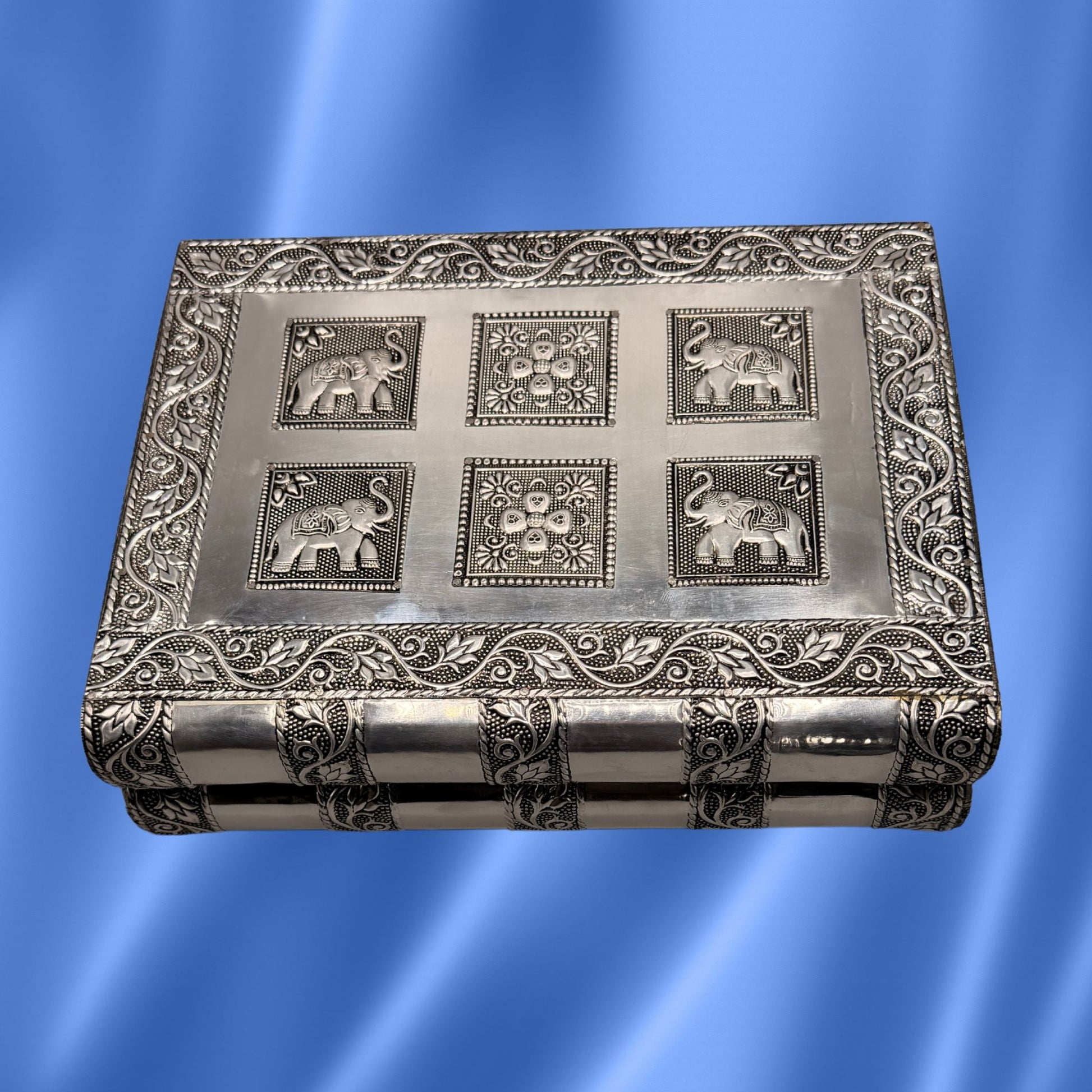 A vintage rectangular silver-toned metal box with embossed floral borders and six raised elephant motifs on the lid, shown against a blue satin-like background.