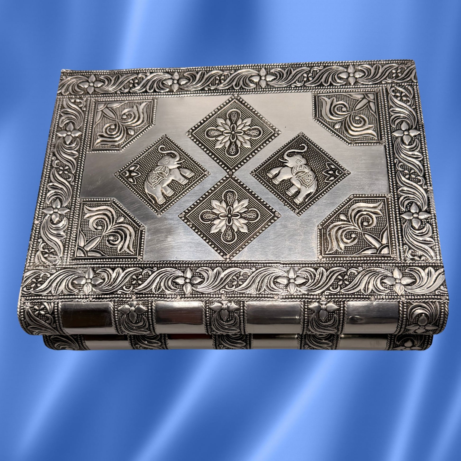 Vintage Indian silver jewelry box with intricate embossed floral borders and elephant motifs in the center, shown against a blue gradient background.