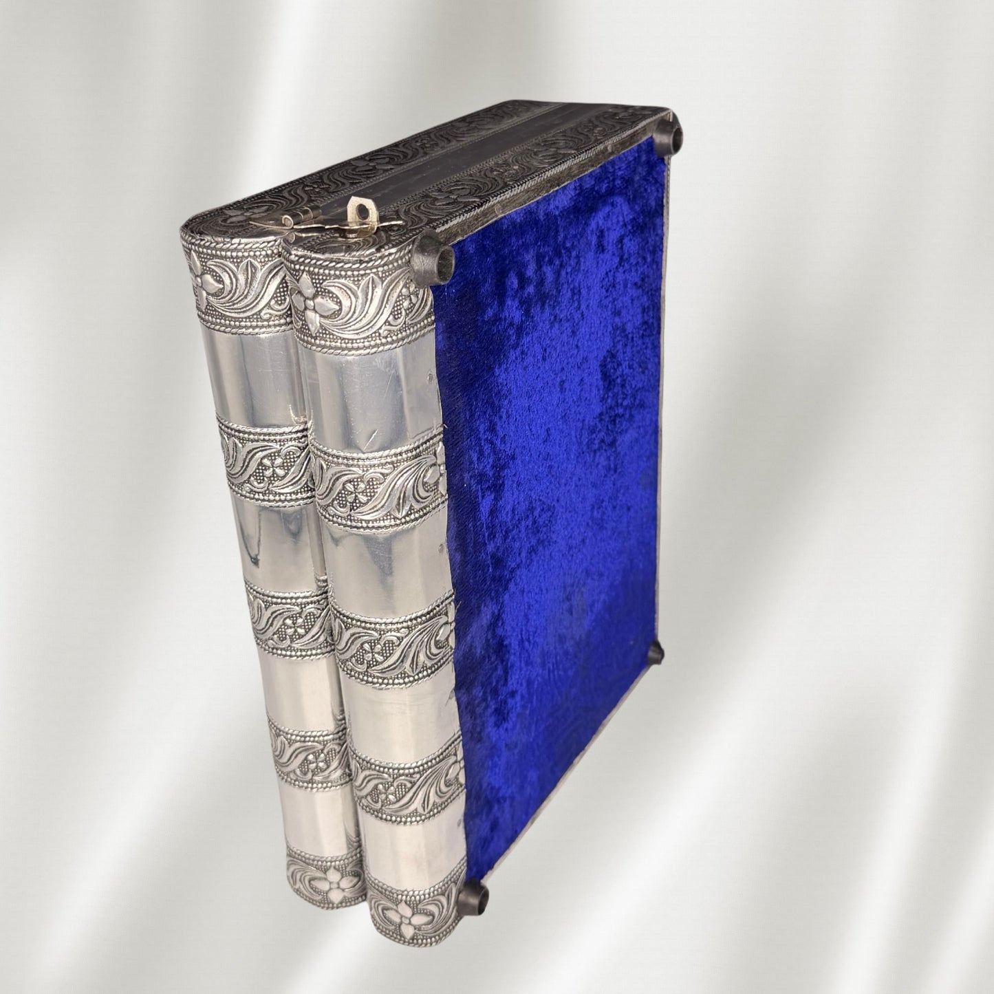 A decorative book-shaped jewelry box with ornate silver-toned embossed detailing on the spine and cover, featuring a rich royal blue velvet bottom cover, set against a draped white fabric background.