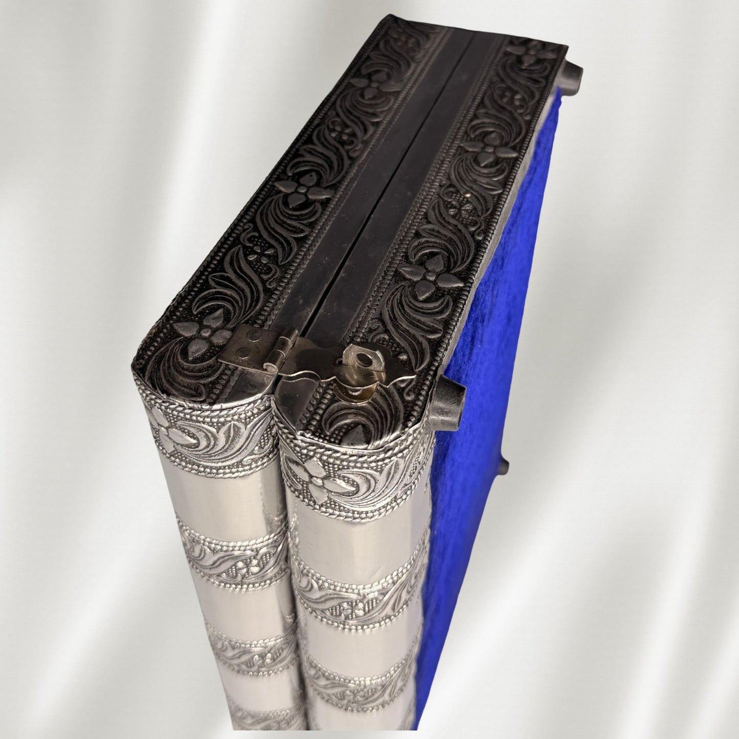 An antique silver jewelry box with intricate floral embossed detailing on the edges shown standing on its side with a white fabric background.