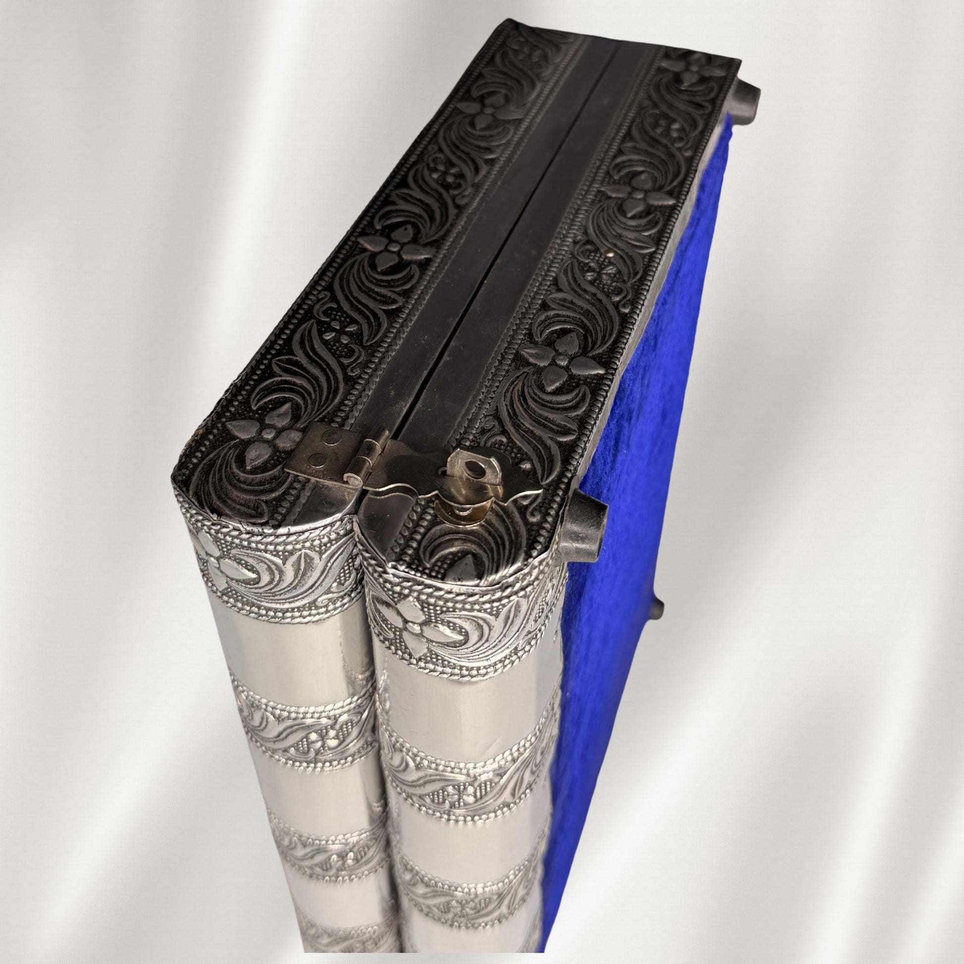 An antique silver jewelry box with intricate floral embossed detailing on the edges shown standing on its side with a white fabric background.