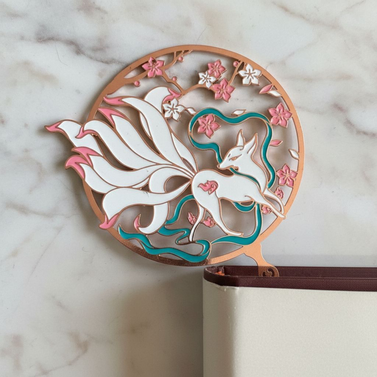 Nine Tailed Brass Fox Bookmark sticking out of the corner of a book against a marble background