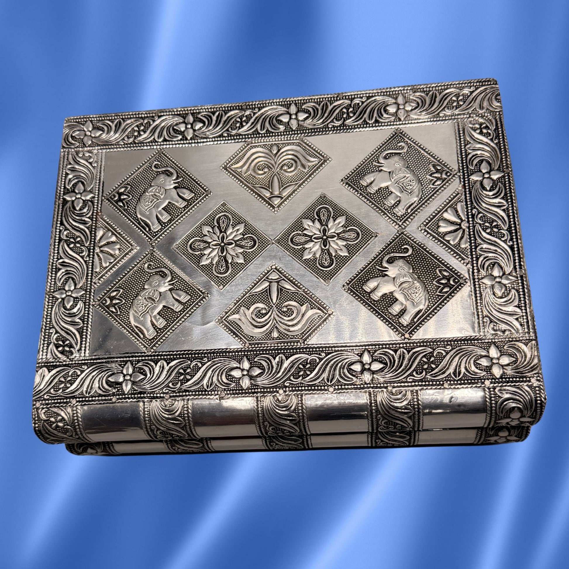 An elephant rectangular silver-toned metal box with intricate embossed floral borders and elephant motifs on the lid, shown against a blue gradient background.