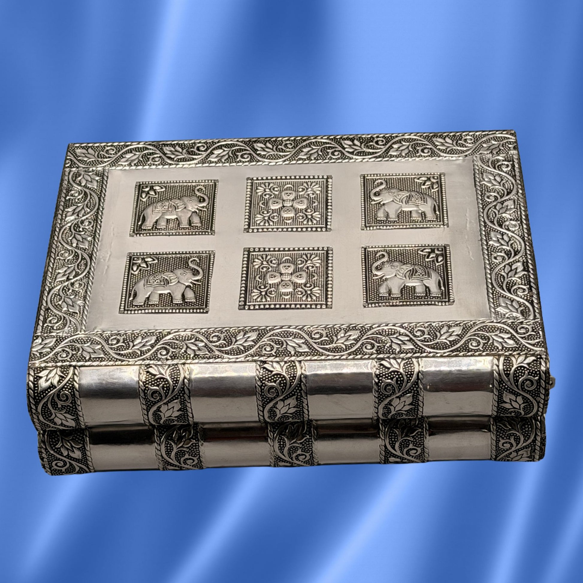 An Indian silver-toned metal jewelry box with embossed floral borders and  elephant motifs on the lid, shown against a blue gradient background.
