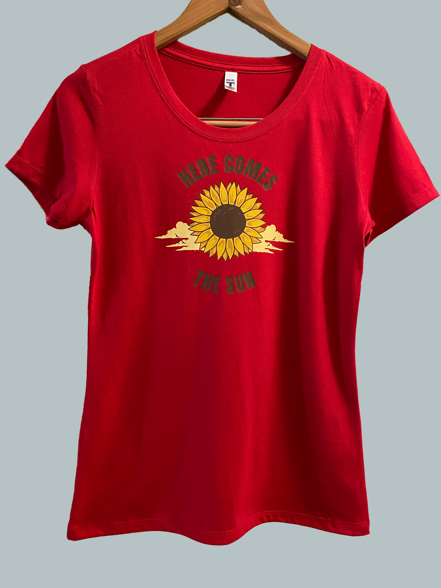 Here Comes The Sun written around a big sunflower that is rising out of the clouds on this red slightly fitted t-shirt Fabric 60% combed ringspun cotton/40% polyester lightweight jersey  Fabric laundered for reduced shrinkage 